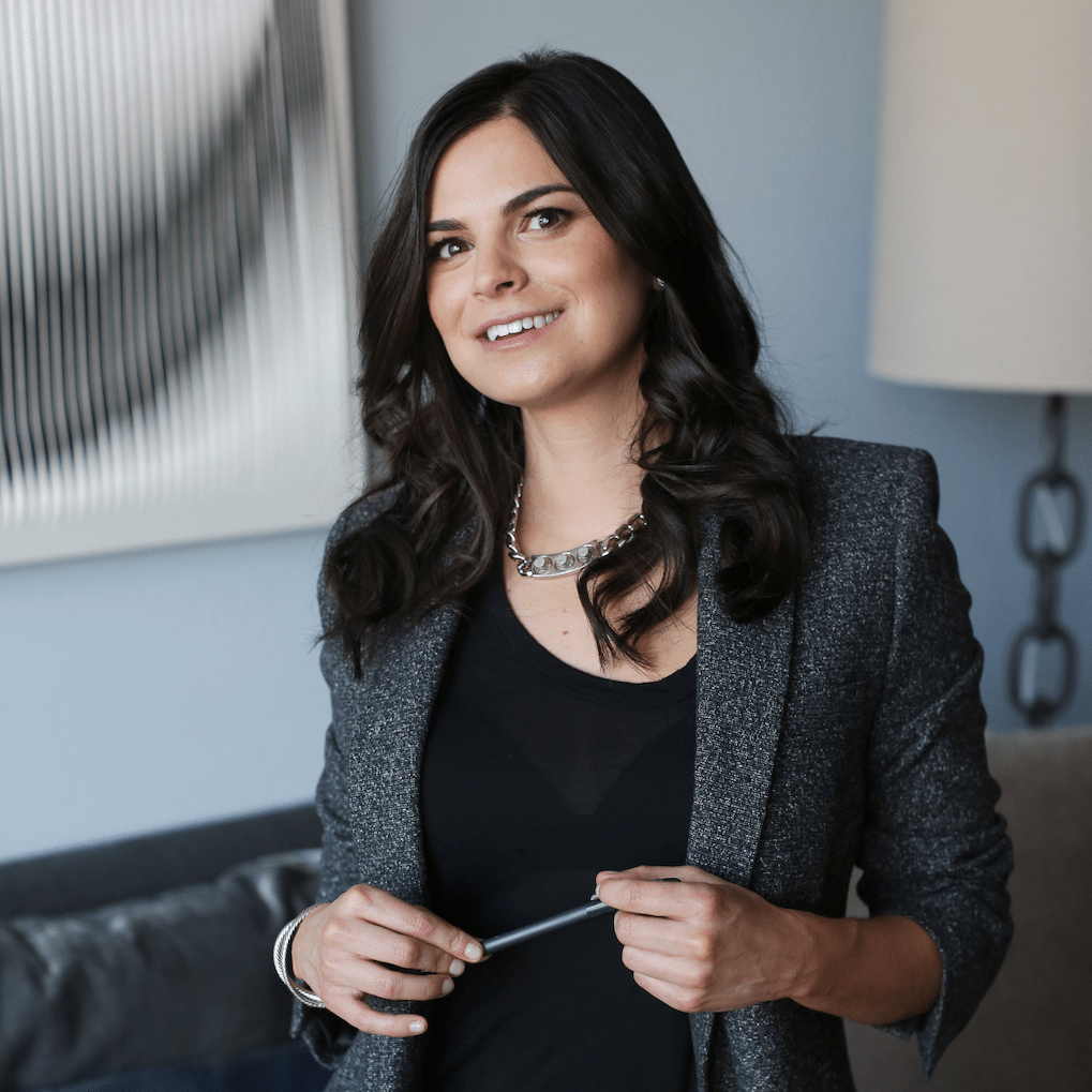 Andrea Serbonich stands in a light-blue office, wearing a grey suite with loosely curled hair