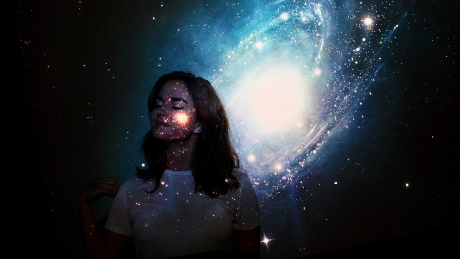 hybrid coaching can combine the intelligence of humans and AI to multiply impact. Image description: a woman stands against a wall with a galaxy projection covering her face and shirt.